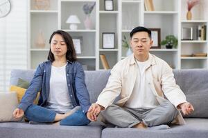 contento asiatico famiglia a casa su divano meditando, coppia uomo e donna seduta nel loto posizione nel vivente camera con chiuso occhi riposo rilassante insieme. foto