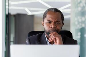 avvicinamento foto. anziano africano americano maschio uomo d'affari, direttore si siede nel il ufficio a il il computer portatile e sembra pensieroso a il schermo. foto