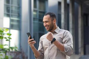 africano americano uomo d'affari al di fuori ufficio edificio utilizzando Telefono, sorridente e contento Tenere mano su trionfo gesto, giovane imprenditore festeggiare vittoria lettura bene investimento notizia in linea foto