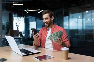 riuscito uomo d'affari nel rosso camicia festeggiare vittoria e bene realizzazione Tenere Telefono e i soldi americano denaro contante dollari nel mani, investitore Lavorando dentro ufficio con il computer portatile avuto reddito. foto