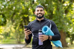 felice, in forma uomo Tenere un' yoga stuoia e acqua bottiglia mentre utilizzando il suo smartphone nel un' soleggiato, verde parco. foto