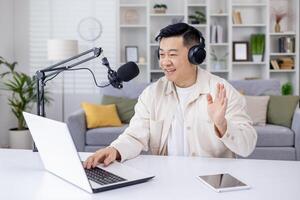 un coinvolgente asiatico uomo agitando mentre registrazione un' Podcast utilizzando un' il computer portatile e microfono impostato su a il suo casa ufficio. foto