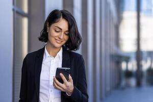 un' giovane bellissimo attività commerciale donna è a piedi al di fuori il ufficio costruzione, un' donna nel attività commerciale Abiti è sorridente, utilizzando un applicazione su il Telefono, navigazione il Internet, digitando un' Messaggio. foto