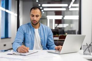 focalizzata indiano uomo d'affari a il suo scrivania nel un' ben illuminato moderno ufficio, Lavorando su un' il computer portatile e scrittura Appunti. foto