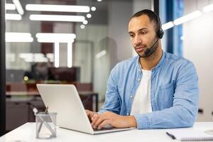 grave giovane musulmano uomo indossare un' cuffia è concentrandosi su Lavorando su un' il computer portatile mentre seduta a un' scrivania nel il ufficio. foto