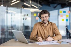 contento maturo ispanico uomo godendo il suo opera con il computer portatile e documenti nel un' moderno ufficio ambientazione. positivo opera ambiente e lavoro soddisfazione concetto. foto