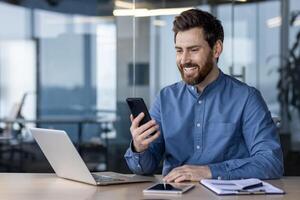 sorridente maschio professionale sms su Telefono mentre Lavorando su il computer portatile a un' contemporaneo ufficio spazio. foto