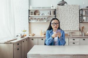 giovane bellissimo asiatico donna nel bicchieri e denim Abiti seduta nel il cucina a casa e utilizzando un' mobile Telefono. quadranti, sorrisi. foto