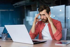 frustrato uomo d'affari depresso a posto di lavoro Lavorando su computer portatile, uomo nel camicia irritato e triste scontento con cattivo opera risultati e realizzazione dentro ufficio. foto