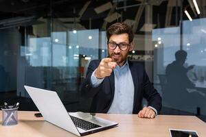 riuscito maturo uomo d'affari guardare a telecamera e puntamento dito inoltrare chiamata per azione, investitore nel attività commerciale completo da uomo dentro moderno ufficio Lavorando su computer portatile, capo con barba e occhiali. foto