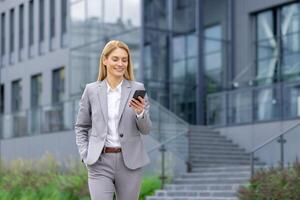 riuscito donna capo passeggiate al di fuori ufficio edificio all'aperto, nel attività commerciale completo da uomo, donna d'affari usi App su Telefono, sorrisi contento legge Internet pagine, sfoglia notizia, scrive testo Messaggio, chiamata. foto