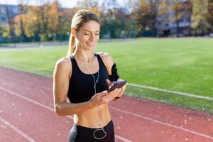 sportivo femmina atleta nel fitness abbigliamento godendo musica mentre interagendo con sua Telefono su un' soleggiato traccia campo. foto