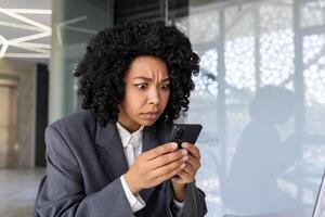 vicino su di africano americano donna lettura cattivo notizia a partire dal televisione, attività commerciale donna avuto rifiuto e fraudolento Azioni in linea, femmina lavoratore dentro ufficio Tenere smartphone irritato lettura. foto