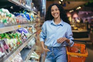 ritratto di contento ispanico donna acquirente nel supermercato, donna con shopping cestino sorridente e guardare a telecamera, vicino scaffali con prodotti nel drogheria Dipartimento foto