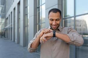 latino americano uomo d'affari nel camicia allegro parlando con amici utilizzando orologio intelligente, uomo al di fuori ufficio edificio utilizzando aggeggio per artificiale intelligenza comandi foto