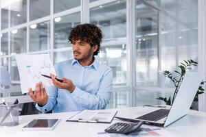 irritato finanziere mostrando in linea rapporto, uomo Lavorando dentro ufficio nervosamente mostrando documento, ispanico uomo nel in linea incontro utilizzando il computer portatile per a distanza comunicazione e chiamata. foto