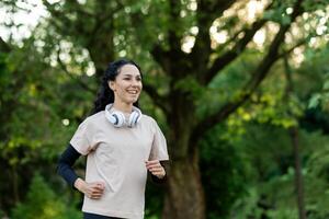 attivo femmina nel maglietta al di sopra di lungo manica con cuffia al di sopra di collo esercizio all'aperto durante giorno nel verde la zona. positivo signora fare jogging nel pubblico parco per conservazione in forma e salutare corpo. foto