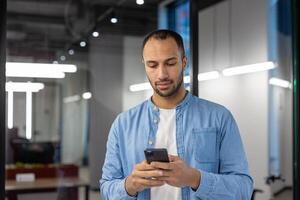 grave giovane ispanico uomo nel denim camicia in piedi nel moderno ufficio e utilizzando mobile Telefono. foto