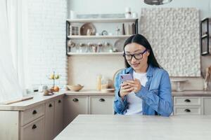 giovane bellissimo asiatico donna digitando Messaggio, donna utilizzando smartphone nel cucina a casa e sorridente foto