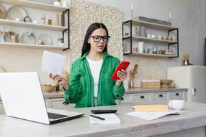 arrabbiato donna seduta a casa utilizzando un' il computer portatile. detiene documenti nel sua mani, tipi su il Telefono foto