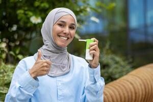 un' allegro musulmano donna nel un' hijab detiene un' gola spray bottiglia e dà un' pollici su su un' soleggiato giorno all'aperto, mostrando Salute e benessere. foto