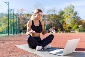 in linea addestramento, femmina allenatore seduta su gli sport stuoia nel stadio a giorno, utilizzando il computer portatile per chiamata per in linea addestramento, attivo stile di vita corsi. foto