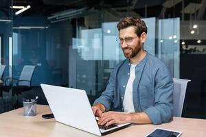 sorridente e allegro uomo d'affari nel casuale camicia Lavorando dentro moderno ufficio, maturo programmatore scrittura codice utilizzando il computer portatile. foto