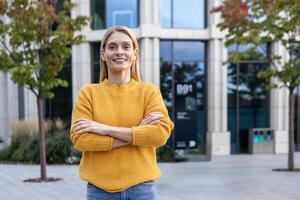 un' professionale donna d'affari sta con fiducia all'aperto, braccia attraversato, indossare un' vivace giallo maglione. lei sorrisi nel un' moderno urbano parco ambientazione. foto