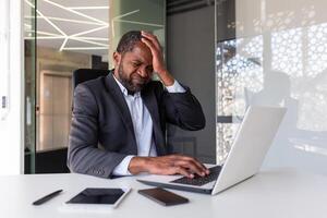 uomo mal di testa dentro ufficio a posto di lavoro, anziano maturo africano americano uomo d'affari oberati di lavoro stressato con opera, capo con il computer portatile seduta a scrivania, Tenere mano su testa. foto