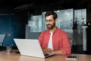 riuscito sorridente uomo Lavorando dentro ufficio con computer portatile, uomo d'affari nel rosso camicia sorridente e digitando su tastiera nel occhiali, programmatore Lavorando Software per programma. foto