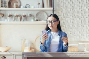 giovane asiatico donna felicemente shopping in linea seduta a casa nel il cucina, donna utilizzando smartphone e banca credito carta per in linea acquisti. foto