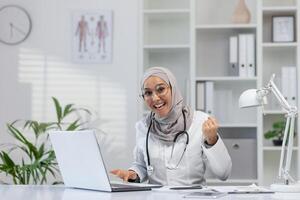 gioioso medio orientale femmina medico nel un' hijab esprimendo eccitazione mentre Lavorando su un' il computer portatile nel un' moderno clinica ufficio. assistenza sanitaria e tecnologia concetto. foto