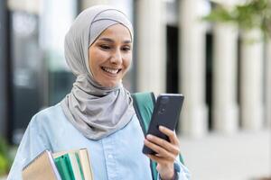 giovane musulmano donna nel hijab a piedi al di fuori Università città universitaria, femmina alunno sorridente contento utilizzando App su Telefono, zaino su indietro e libri nel mani. foto