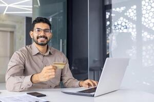 un' allegro giovane uomo nel un' inteligente camicia fabbricazione un in linea transazione con un' credito carta su un' il computer portatile nel un' moderno ufficio ambientazione. foto