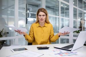 ritratto di frustrato e irritato femmina finanziere dentro ufficio a posto di lavoro, attività commerciale donna guardare scontento a telecamera Tenere finanziario rapporti e contratti con grafici e grafici nel mani. foto