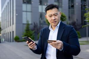 avvicinamento foto di un' preoccupato giovane asiatico uomo seduta su un' panchina vicino un ufficio edificio e guardare confusamente a il Telefono, e credito carta nel il suo mani.