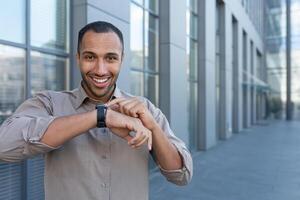 latino americano uomo d'affari nel camicia allegro parlando con amici utilizzando orologio intelligente, uomo al di fuori ufficio edificio utilizzando aggeggio per artificiale intelligenza comandi foto