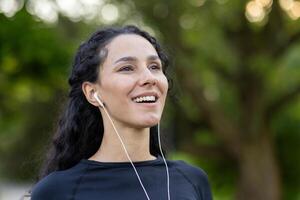 giovane bellissimo donna durante attivo fisico esercizio nel il parco, riposa e respira fresco aria, ispanico donna nel cuffie usi un App su sua Telefono per ascolta per musica e in linea podcast. foto