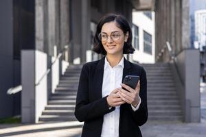 sorridente giovane donna nel un' attività commerciale completo da uomo al di fuori un ufficio centro, Tenere un' Telefono nel sua mani e guardare imbronciato per il lato. foto