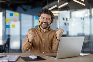 allegro maschio professionale esultante trionfo con il computer portatile nel un' moderno ufficio ambientazione, mostrando positivo emozioni e successo. foto