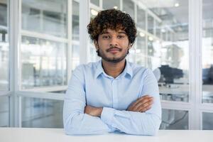 ritratto di un' fiducioso giovane uomo con Riccio capelli, vestito nel un' leggero blu camicia, seduta nel un' ben illuminato ufficio ambiente. braccia attraversato, lui trasuda professionalità e avvicinabilità. foto