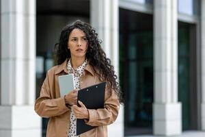 un' professionale donna sta al di fuori un' moderno costruzione, in bilico con un' tavoletta e appunti nel mano. sua alla moda trincea cappotto e inteligente abbigliamento suggerire attività commerciale e efficienza nel un urbano ambientazione. foto