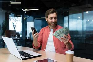 riuscito uomo d'affari nel rosso camicia festeggiare vittoria e bene realizzazione Tenere Telefono e i soldi americano denaro contante dollari nel mani, investitore Lavorando dentro ufficio con il computer portatile avuto reddito. foto