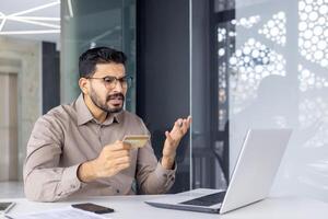 indiano uomo d'affari nel un' moderno ufficio, confuso mentre guardare a un' credito carta, con un' il computer portatile Aperto nel davanti di lui. esprime perplessità e finanziario preoccupazione. foto