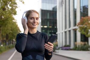 un' giovane adulto donna indossare atletico Ingranaggio ascolta per musica su sua senza fili cuffie mentre Tenere un' smartphone, evocando un' senso di tempo libero e tecnologia nel un urbano ambientazione. foto