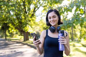 ritratto di un' giovane femmina atleta e allenatore in piedi nel il parco indossare cuffia, Tenere un' Telefono e un' bottiglia di acqua nel sua mani, guardare a il telecamera con un' Sorridi. foto