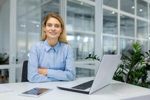 fiducioso femmina professionale sorridente a posto di lavoro con computer portatile, tavoletta, e interno pianta nel un' contemporaneo ufficio ambientazione. foto