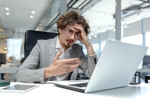 giovane maschio professionale con Riccio capelli Spettacoli frustrazione mentre Lavorando su il computer portatile nel un' moderno ufficio ambiente. foto