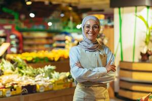 ritratto donna nel un' hijab come un' venditore nel un' supermercato negozio, un' commessa con braccia attraversato sorrisi e sembra a il telecamera, un grembiule vende mele e frutta, un' musulmano donna è soddisfatto nel bicchieri foto