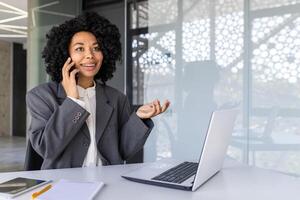 riuscito donna d'affari parlando su il Telefono dentro il ufficio a posto di lavoro, africano americano donna sorridente contento Lavorando utilizzando il computer portatile a opera. foto